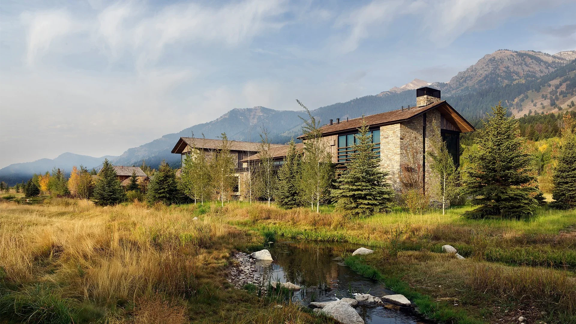 A modern mountain house with stone walls, large windows, and a chimney, surrounded by trees and grass, with mountains in the background and a small creek in the foreground.
