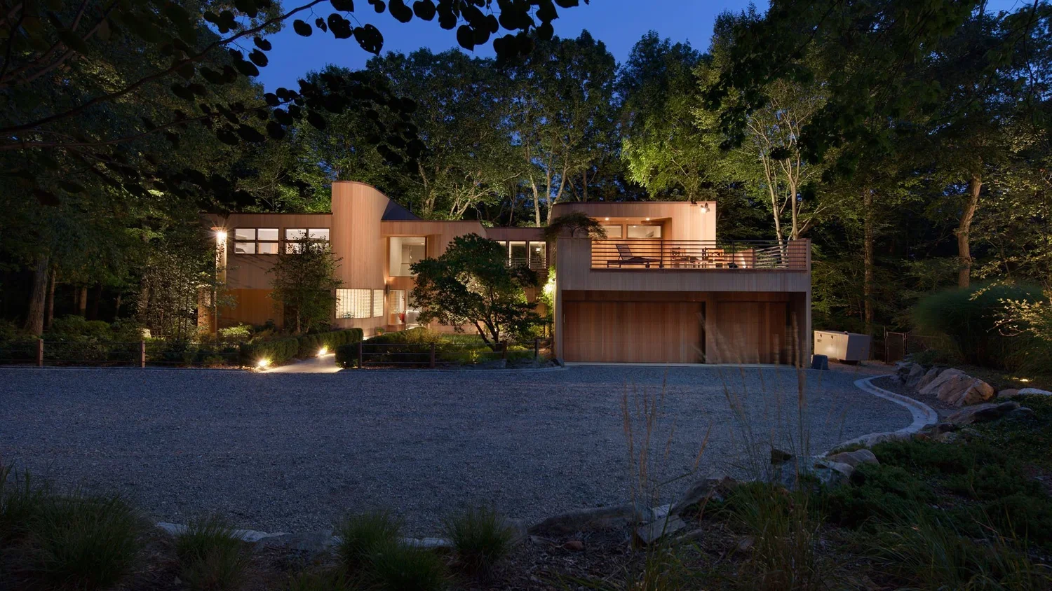 Modern house with wooden exterior illuminated at night, surrounded by trees and greenery, with a gravel driveway in the foreground.