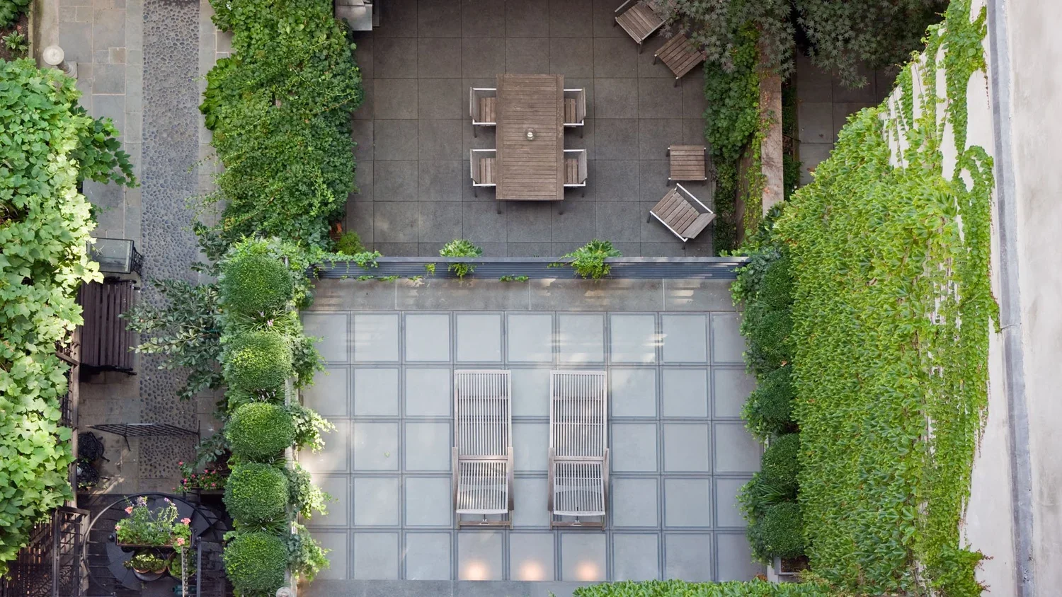 Top-down view of a garden terrace with a central open space containing two lounge chairs, surrounding greenery including trimmed bushes and climbing plants on a wall, and a small seating area with a table and chairs on the left.