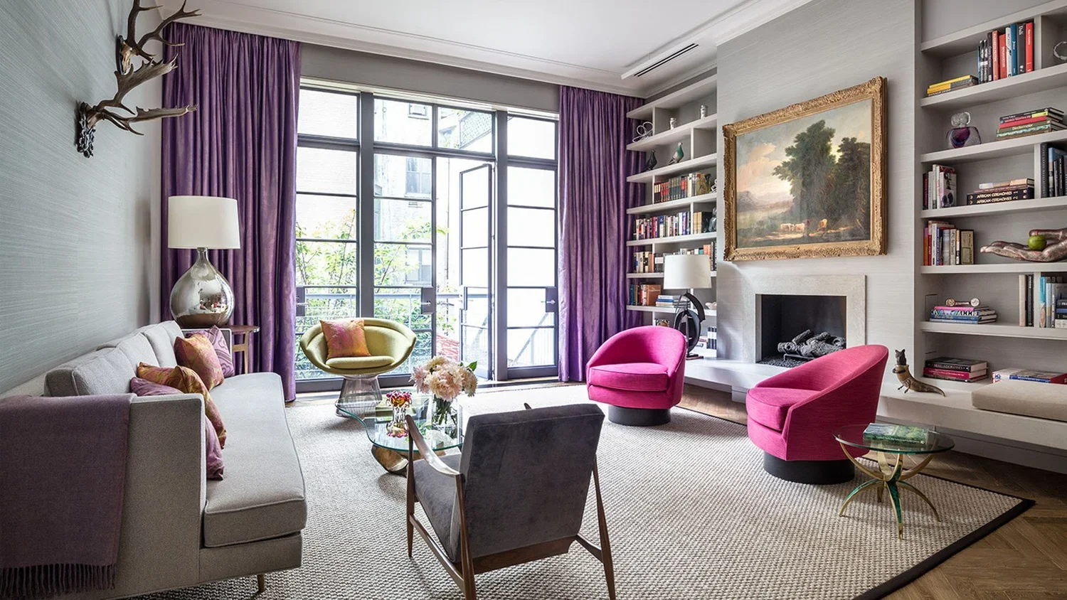 Living room with purple curtains, a gray sofa with colorful pillows, yellow and pink chairs, a glass coffee table with flowers, built-in bookshelves, a fireplace, a large landscape painting, and natural light from glass doors leading outside.