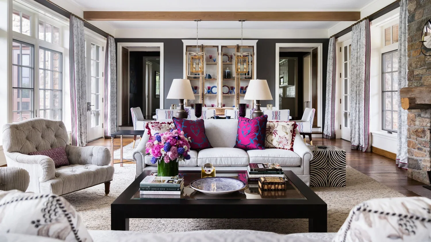 A living room with a beige armchair on the left, a white sofa with colorful pillows in the center, and a black coffee table with books and a floral arrangement. Behind the sofa are shelves with decorative items, and two table lamps. The room has larg