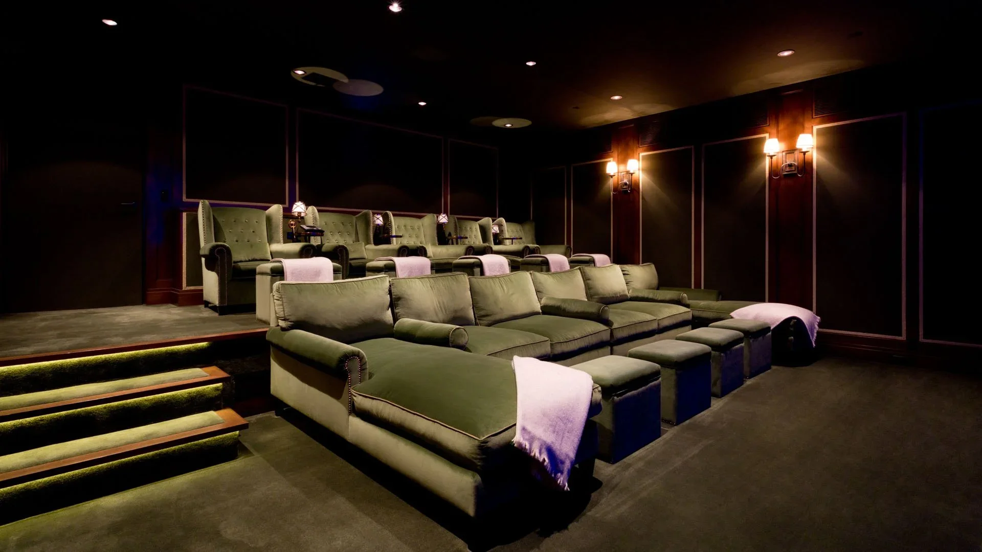 A luxury home theater room with green velvet recliner chairs in the background, a green sofa with matching pillows in the foreground, and dark wood paneled walls with wall sconces providing warm lighting.
