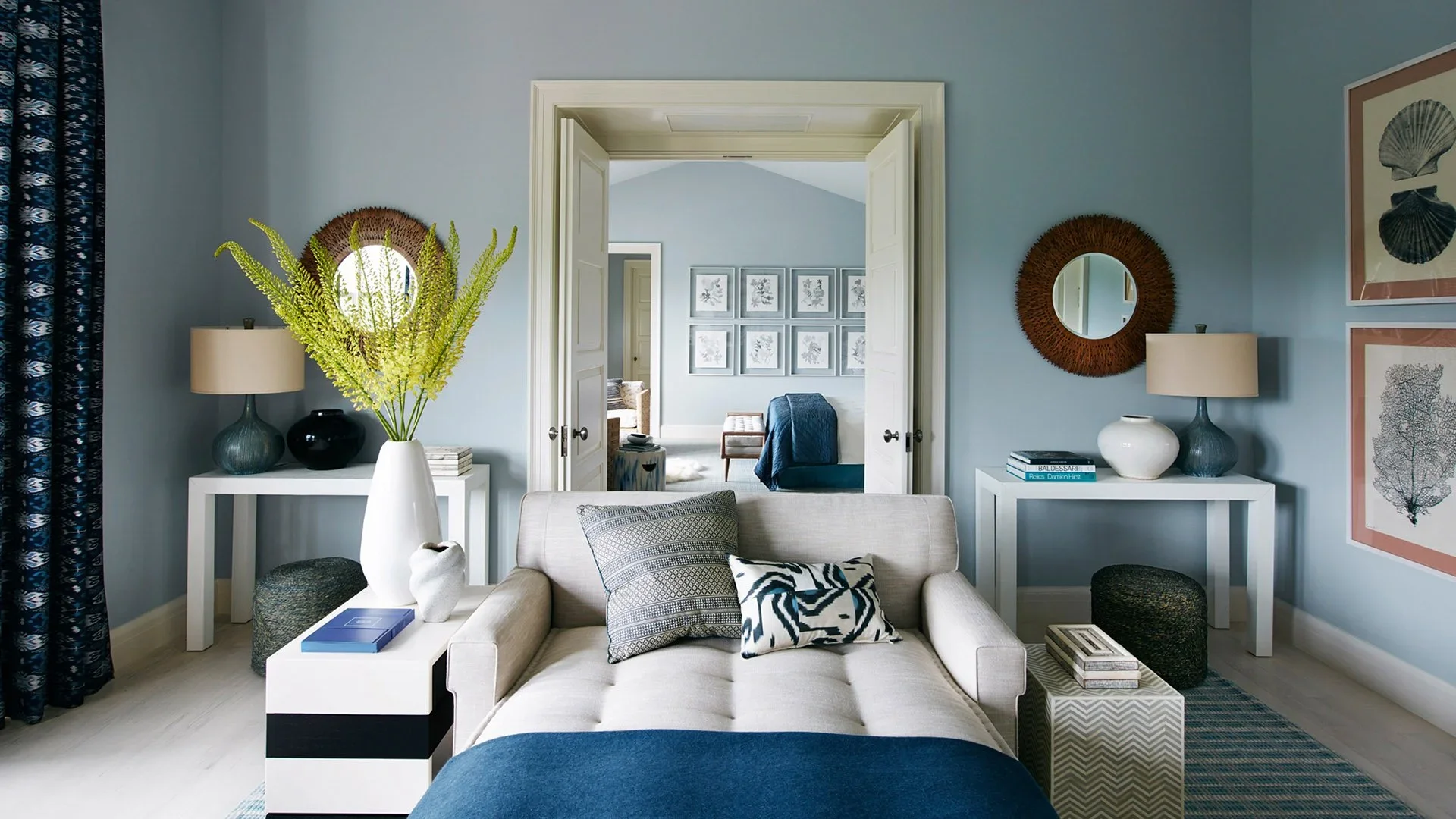 Living room with beige sofa, decorative pillows, white tables with vases, lamps, books, large yellow-green floral arrangement, and wall art, leading to a bedroom with framed botanical prints.