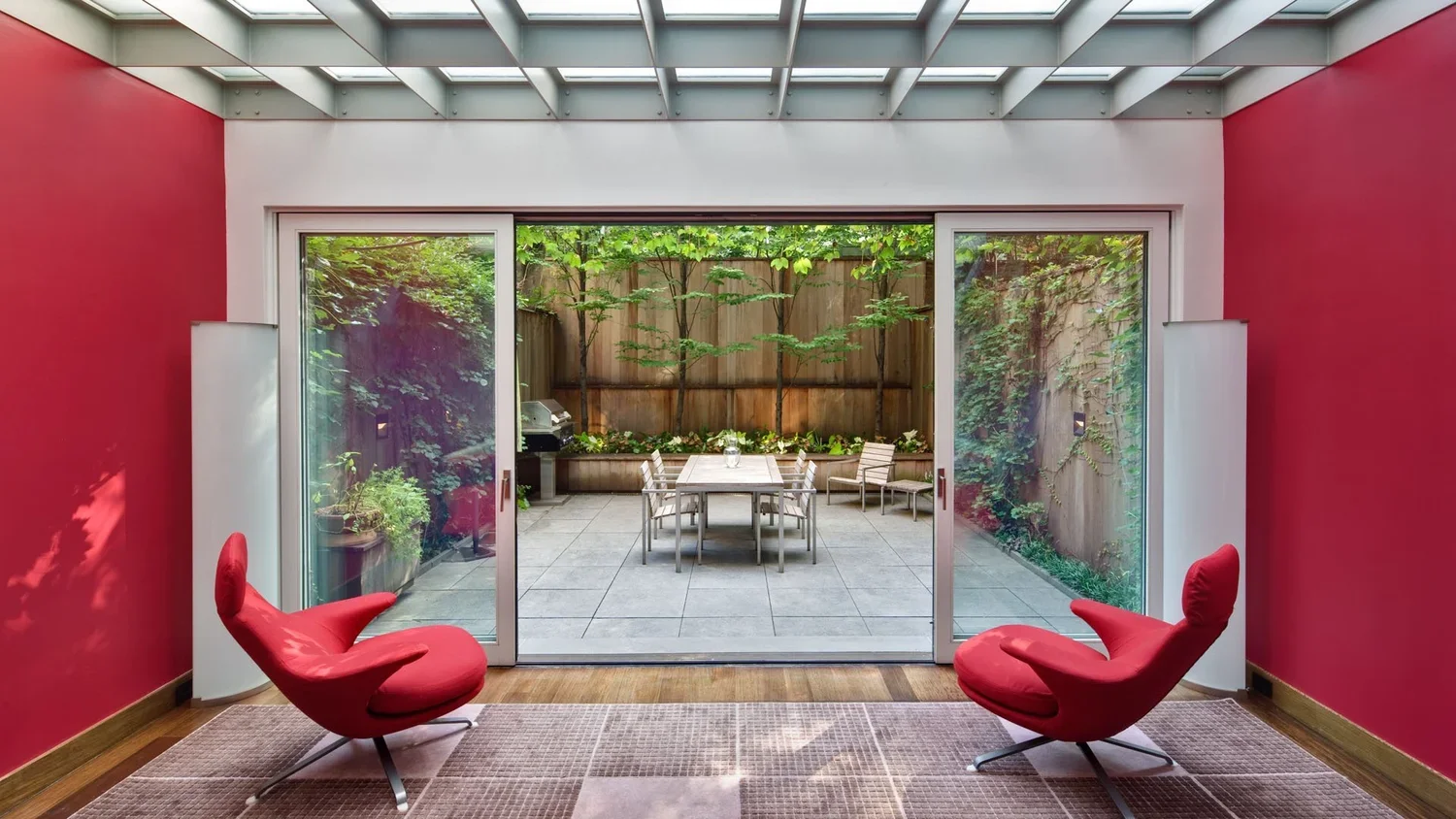 Interior room with pink walls, red modern lounge chairs, and a sliding glass door leading to a small patio with trees, a table, chairs, and a grill.