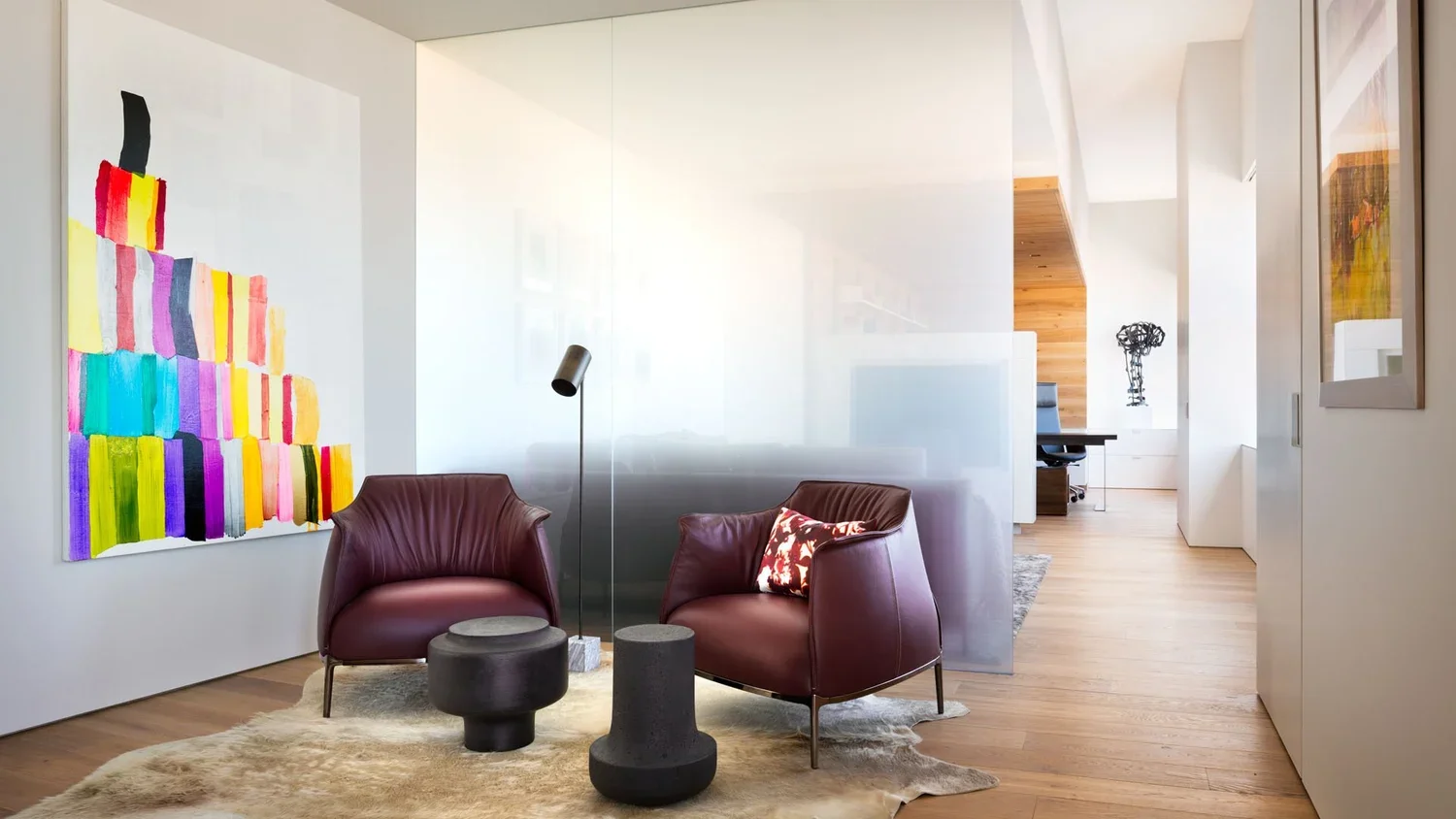 Modern living room with two purple armchairs, a beige carpet, abstract wall art, and wooden flooring. Background includes a glass partition, a desk, and contemporary sculptures.