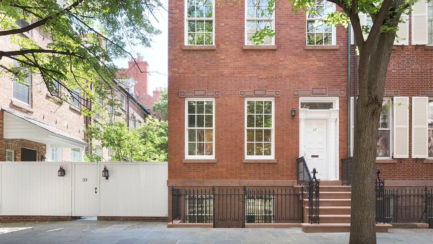A row of brick townhomes with white window frames and front steps, trees with green leaves, and a white fence.