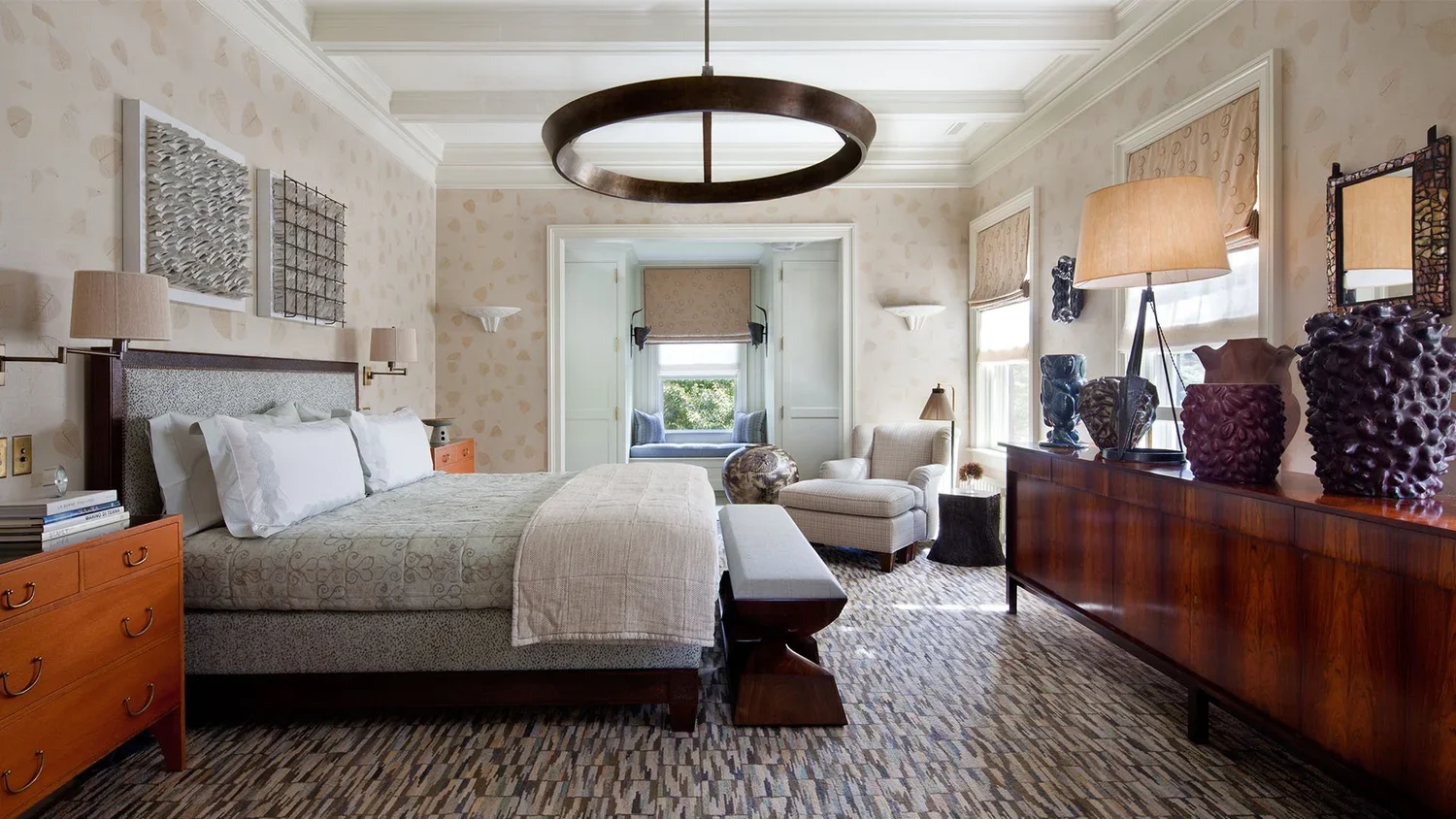 A well-decorated hotel bedroom with a bed, nightstands, artwork, lamps, a sitting nook by the window, and decorative vases and sculptures on a sideboard.