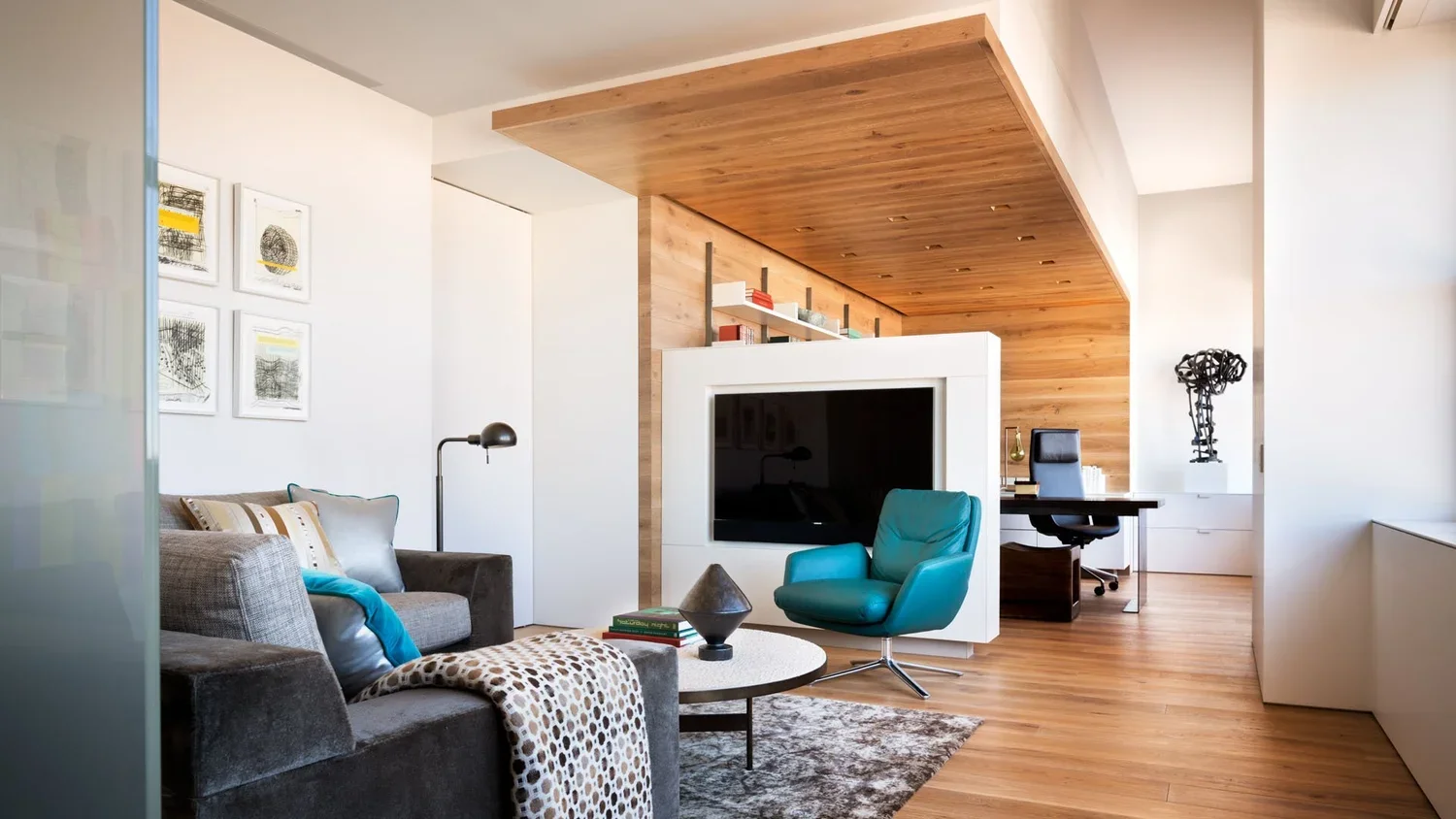 Modern living room with gray sofa, colorful pillows, a teal lounge chair, a round coffee table with books and decor, and a wooden wall with a TV and a workspace area in the background.