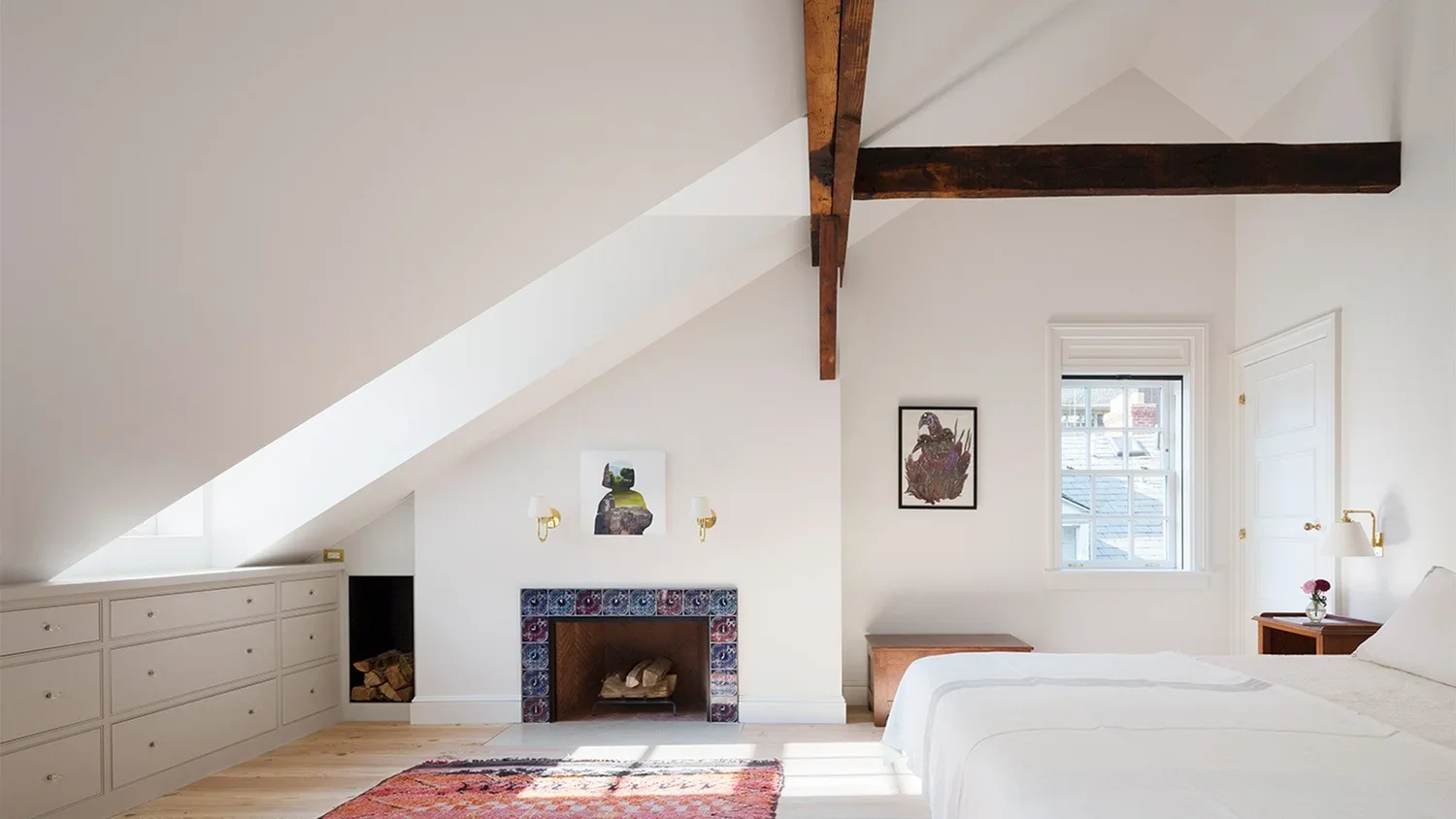 Bright bedroom with white walls, sloped ceiling, and exposed wooden beams. A fireplace with decorative tiles is centered on the back wall, with artwork above it. A window to the right lets in natural light, and there is a bed with white linens on the