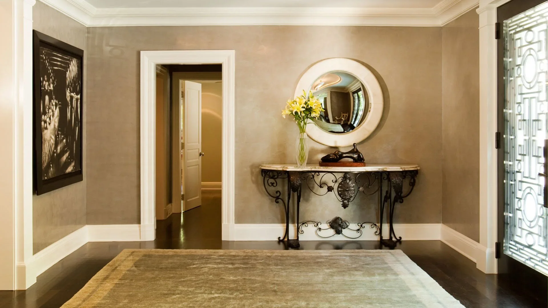 Elegant entryway with beige walls, dark hardwood floors, white trim, and decorative black iron console table with a marble top. A tall, clear vase with yellow lilies and a black decorative object sit on the table. A round mirror hangs above the table