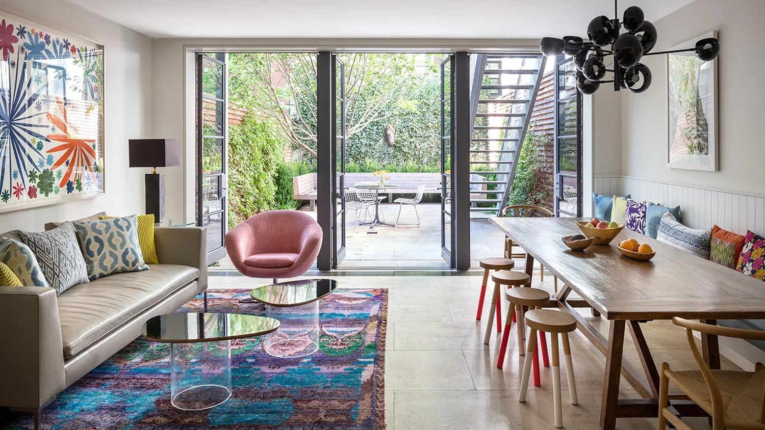 Living room with a cream sofa with patterned pillows, pink chair, glass front doors opening to greenery patio, wooden dining table with stools, colorful artwork and modern black chandelier.