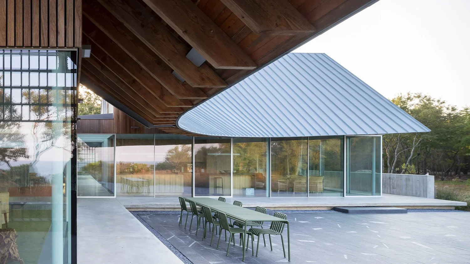 Modern building with a wooden overhang, glass walls, and a metal-paneled roof. An outdoor patio with a long table and green chairs are in the foreground, with trees and water in the background.