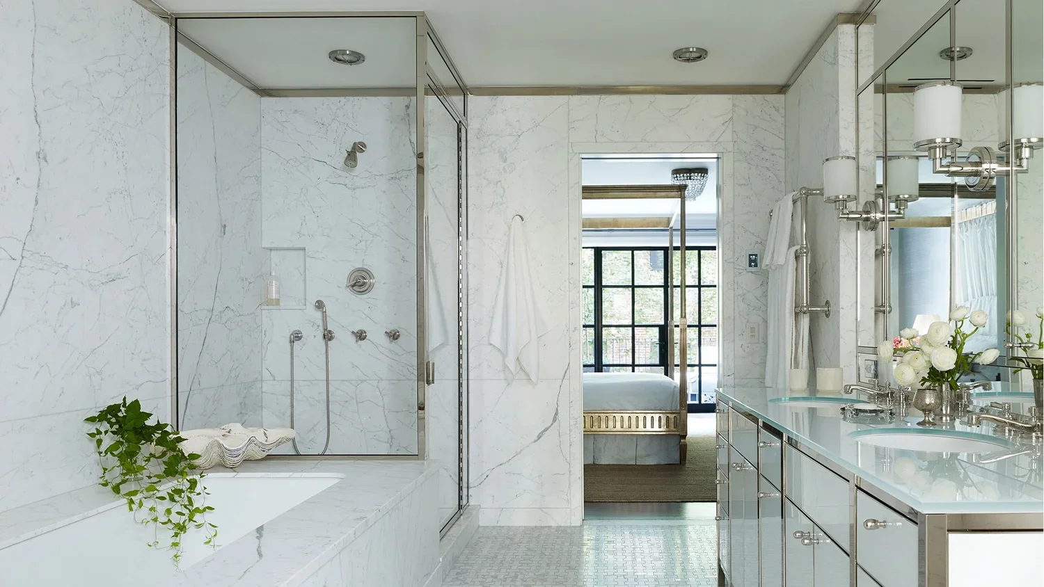 Modern bathroom with white marble walls, a glass shower enclosure, a bathtub with a green plant, and a long vanity with double sinks, flowers, and large mirrors.