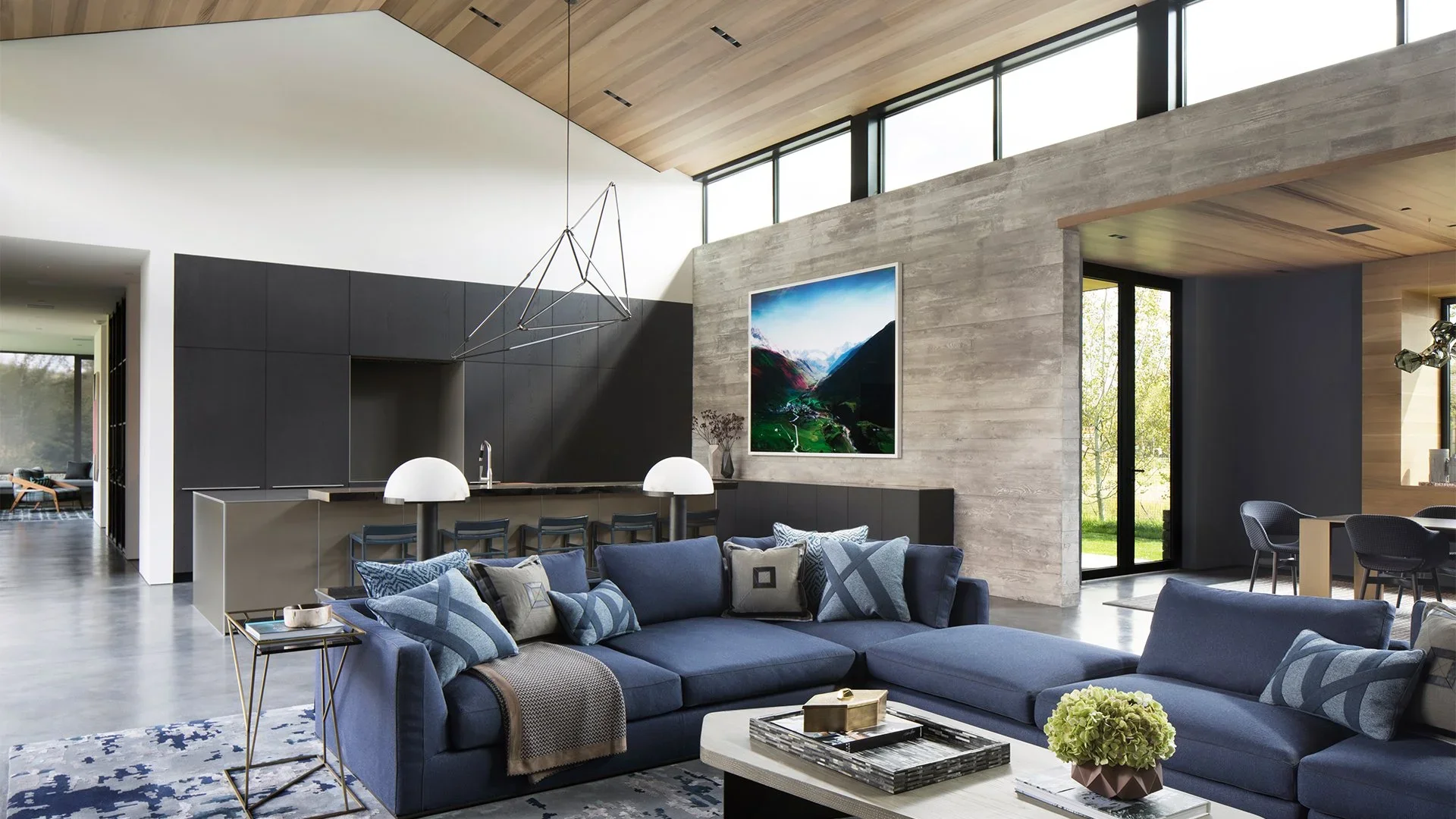 Modern living room with blue sectional sofa, white coffee table, and gray throw blanket. Kitchen area with black cabinetry and island with bar stools. Large window and wooden ceiling with geometric pendant light.
