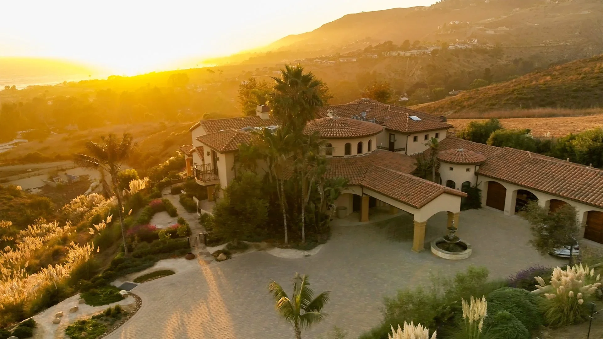 Elegant Mediterranean-style house with red tiled roof, surrounded by lush landscaping and palm trees, near a scenic hillside at sunset.