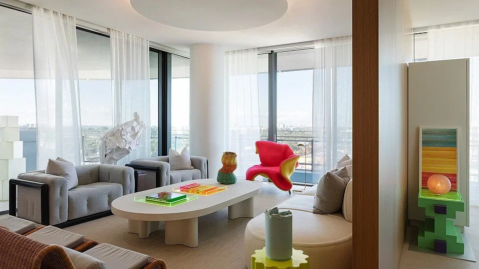 Modern living room with large windows, gray armchairs, a white coffee table, a red and yellow lounge chair, decorative objects, and a city view.