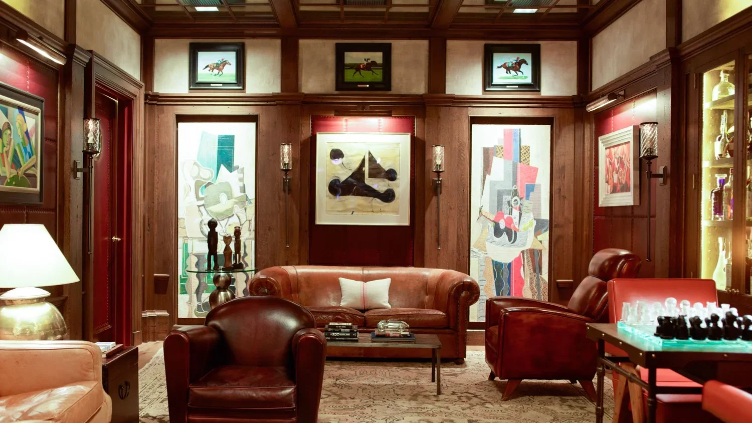 A cozy living room with wooden walls, leather armchairs, a sofa with a pillow, modern art paintings on the walls, a chess game on a table, and dim lighting.