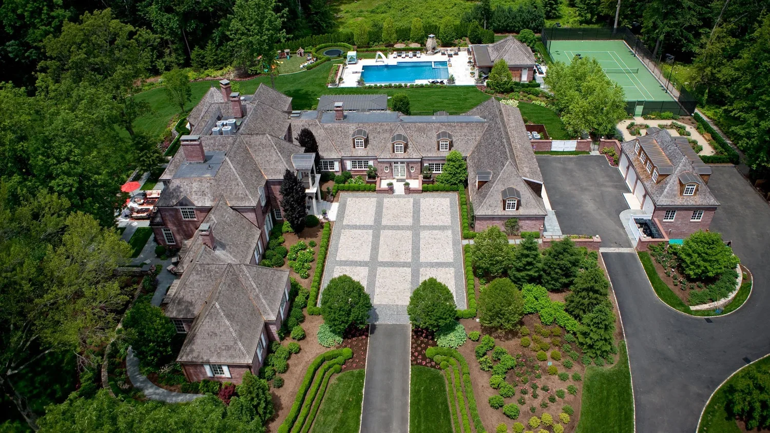 Aerial view of a large mansion with a paved courtyard, surrounded by manicured gardens, trees, a swimming pool, tennis courts, and lush greenery.