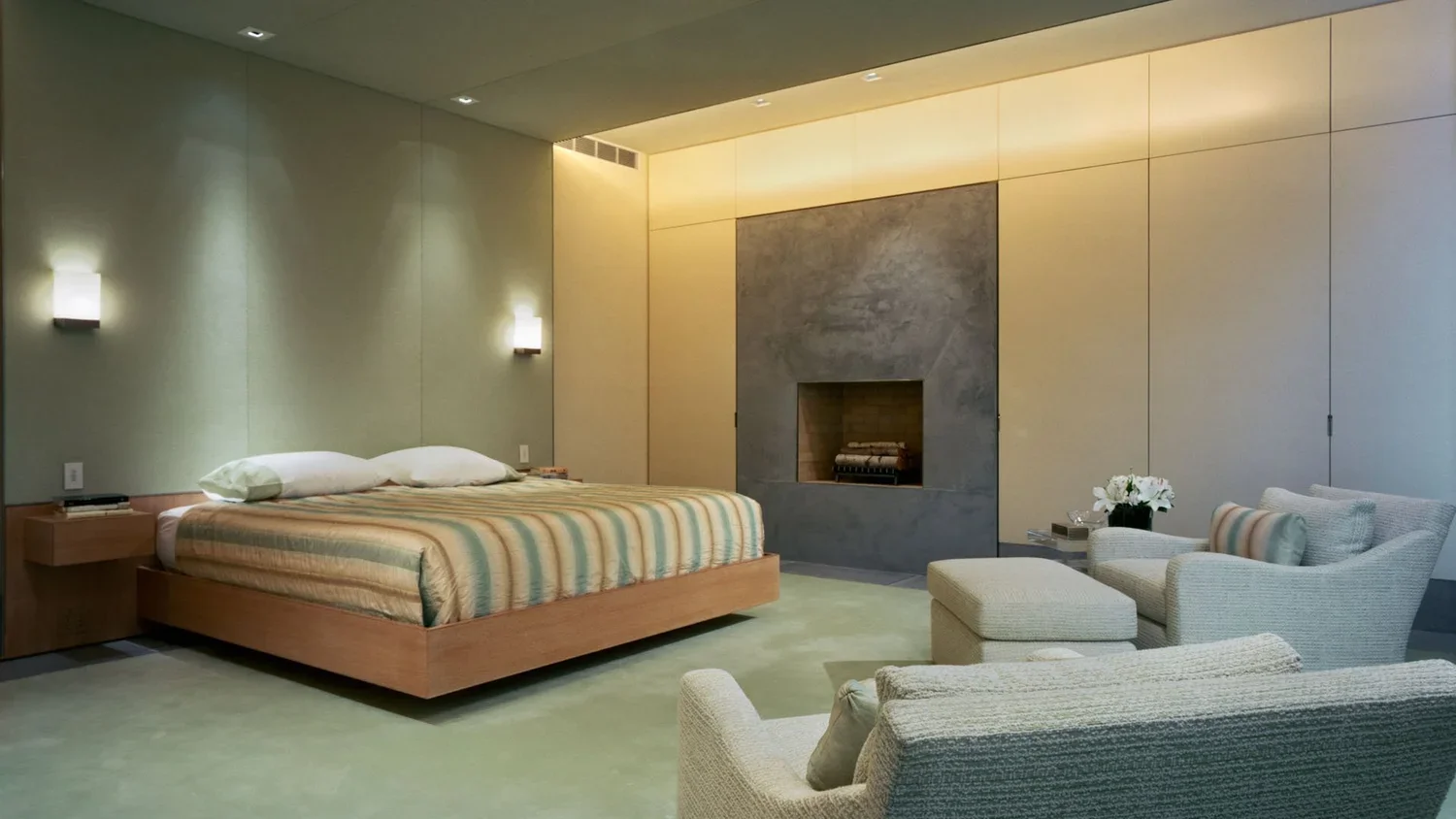 A bedroom with a large wooden bed, two pillows, and colorful striped bedding. Next to the bed are small wooden nightstands with books and a clock. A modern fireplace with a gray surround is on the wall opposite the bed. To the right of the fireplace,