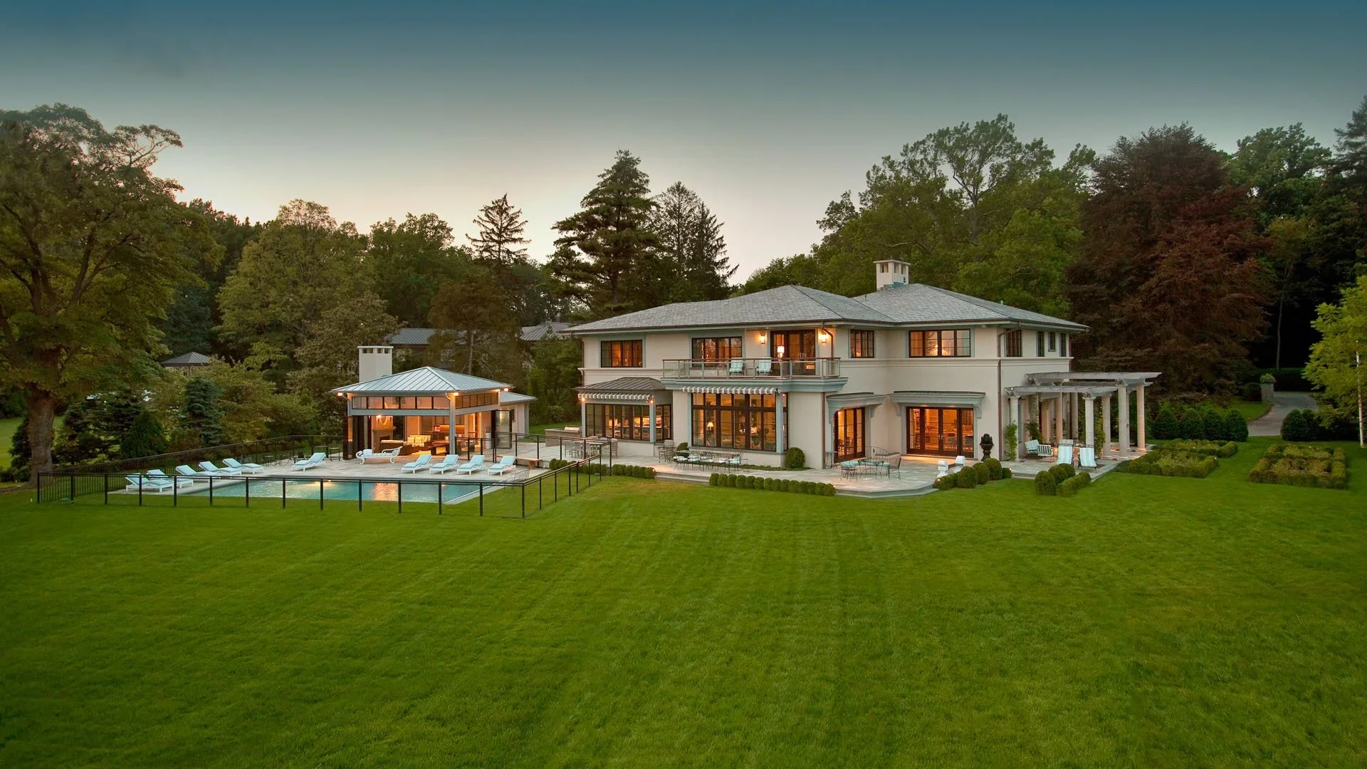 Large luxury house with a swimming pool and a pool house, surrounded by green lawn and tall trees at dusk.
