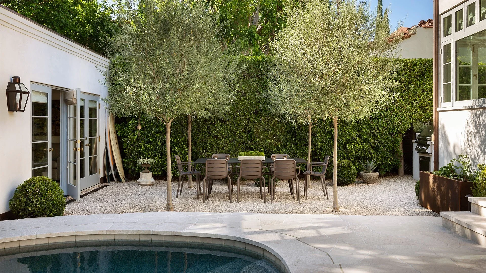 Outdoor patio with a swimming pool in the foreground, a dining table with chairs, trees, and a hedge in the background, and part of a house with large windows and an outdoor grill.