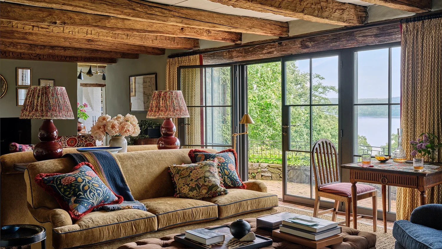 Living room with a yellow sofa, decorative pillows, floral lamps, a small table with books, and glassware, facing large glass doors opening to a balcony overlooking a lakeside view with trees and water.