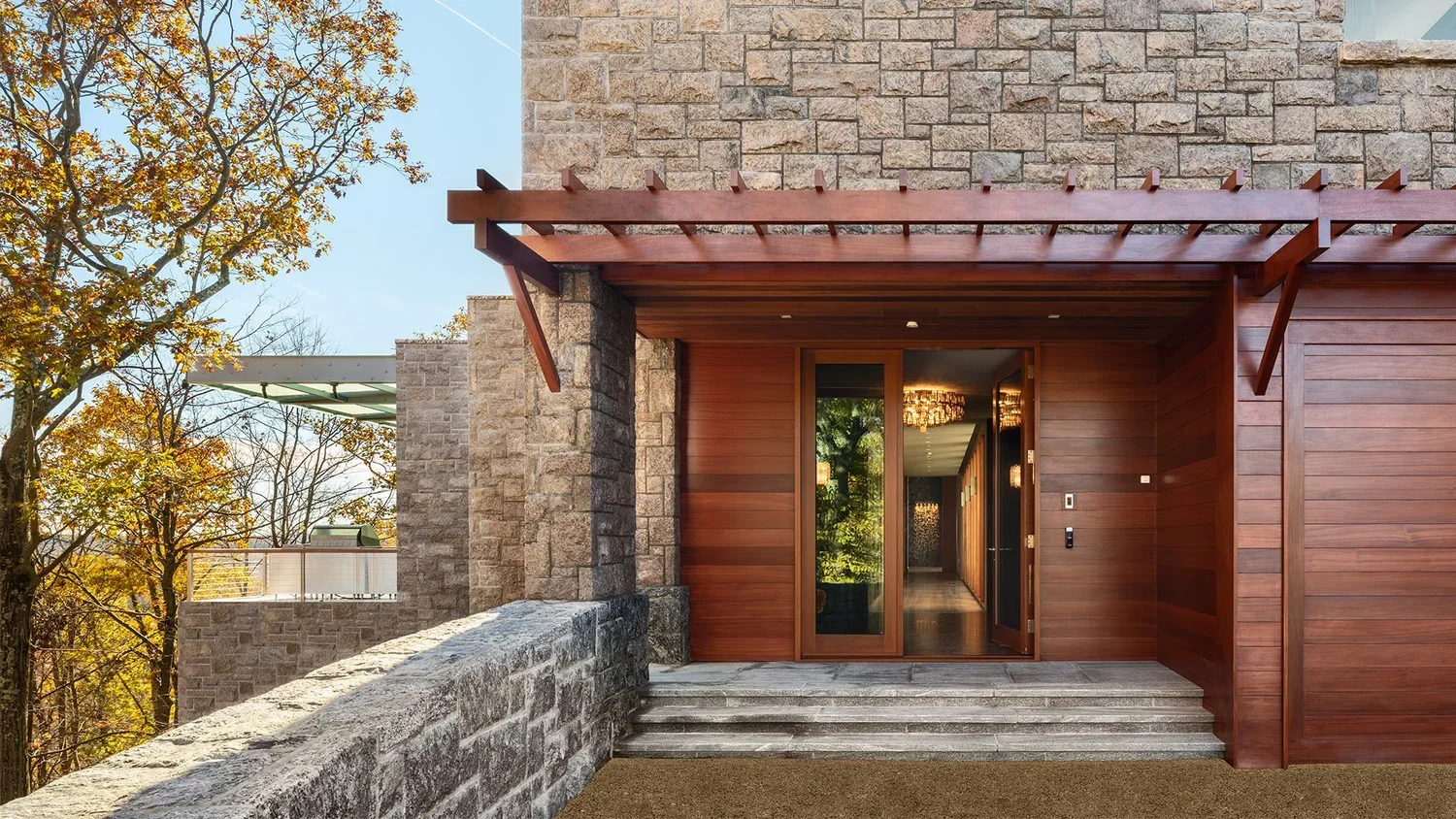 Exterior view of a modern house entrance with wooden paneling, stone walls, glass sliding door, a set of three stone steps leading up to the door, and a wooden pergola overhead during daytime, with autumn trees in the background.