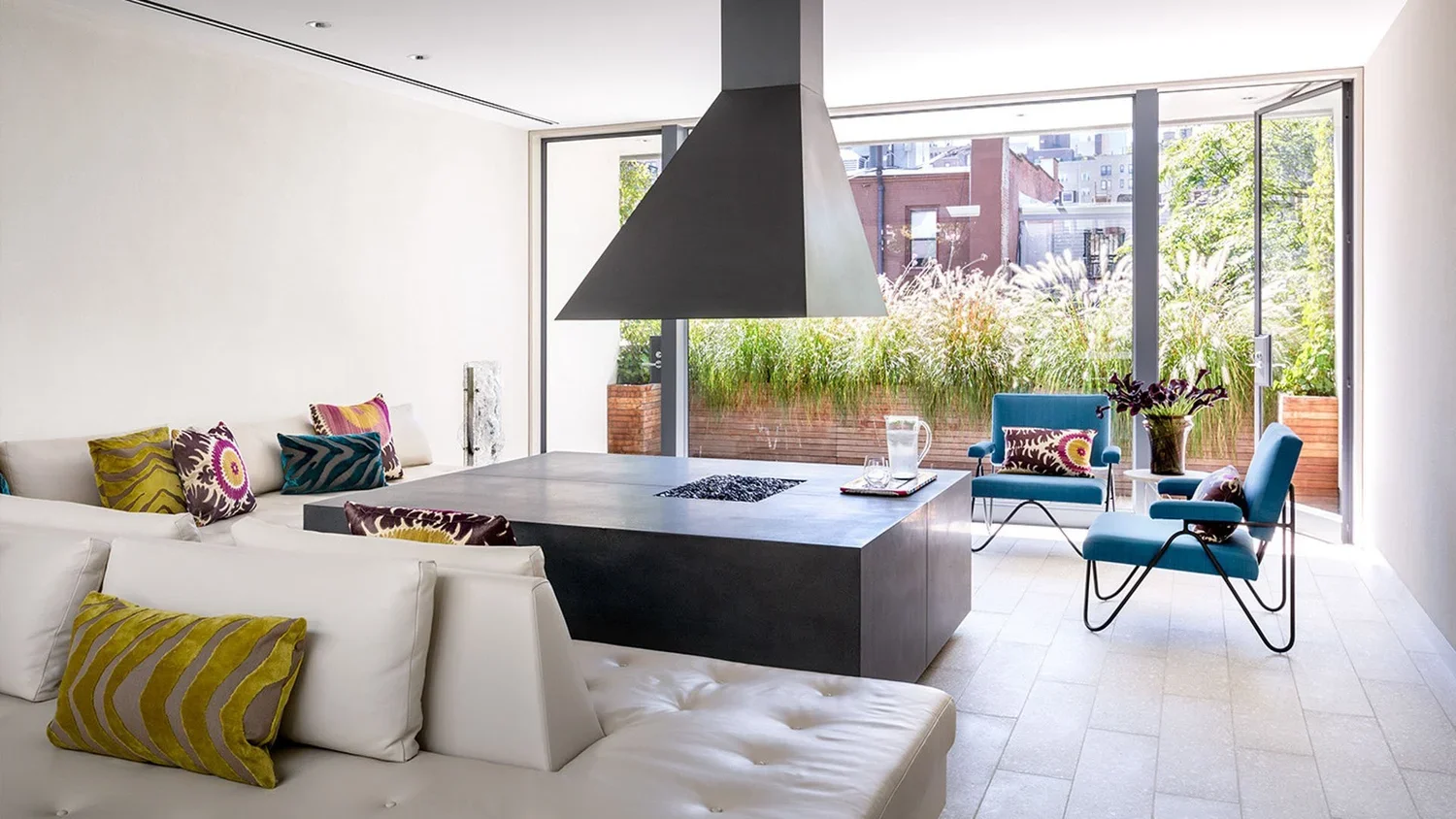 Modern living room with white sofa, colorful throw pillows, two blue chairs, large glass sliding doors, plants outside, and a black fireplace on a dark gray table.
