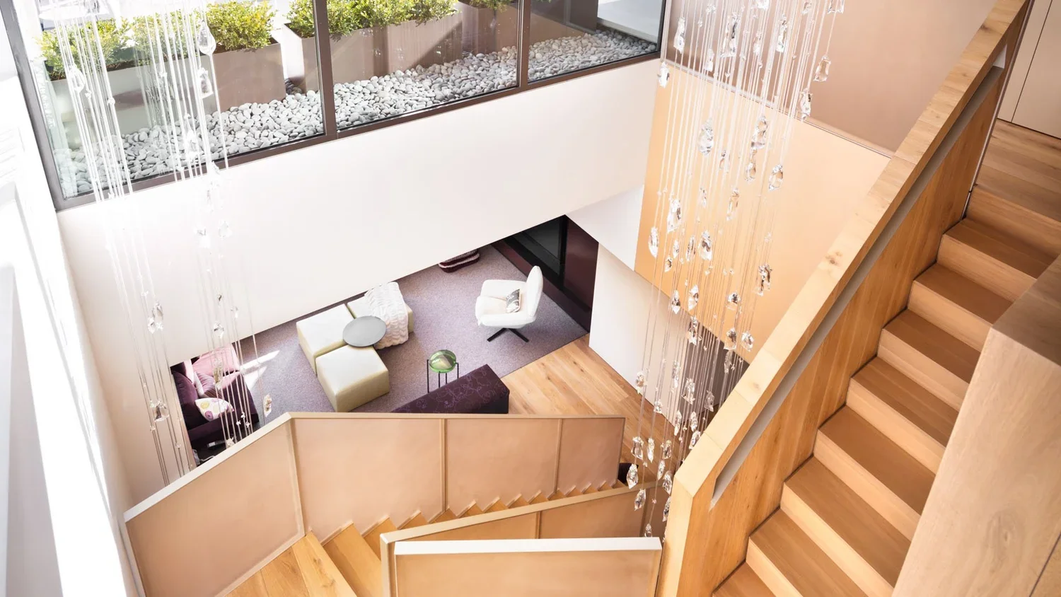 Interior view of a modern multi-level house with wooden stairs, hanging crystal chandeliers, and a glass wall overlooking plants and rocks outside; focusing on the staircase and upper level.