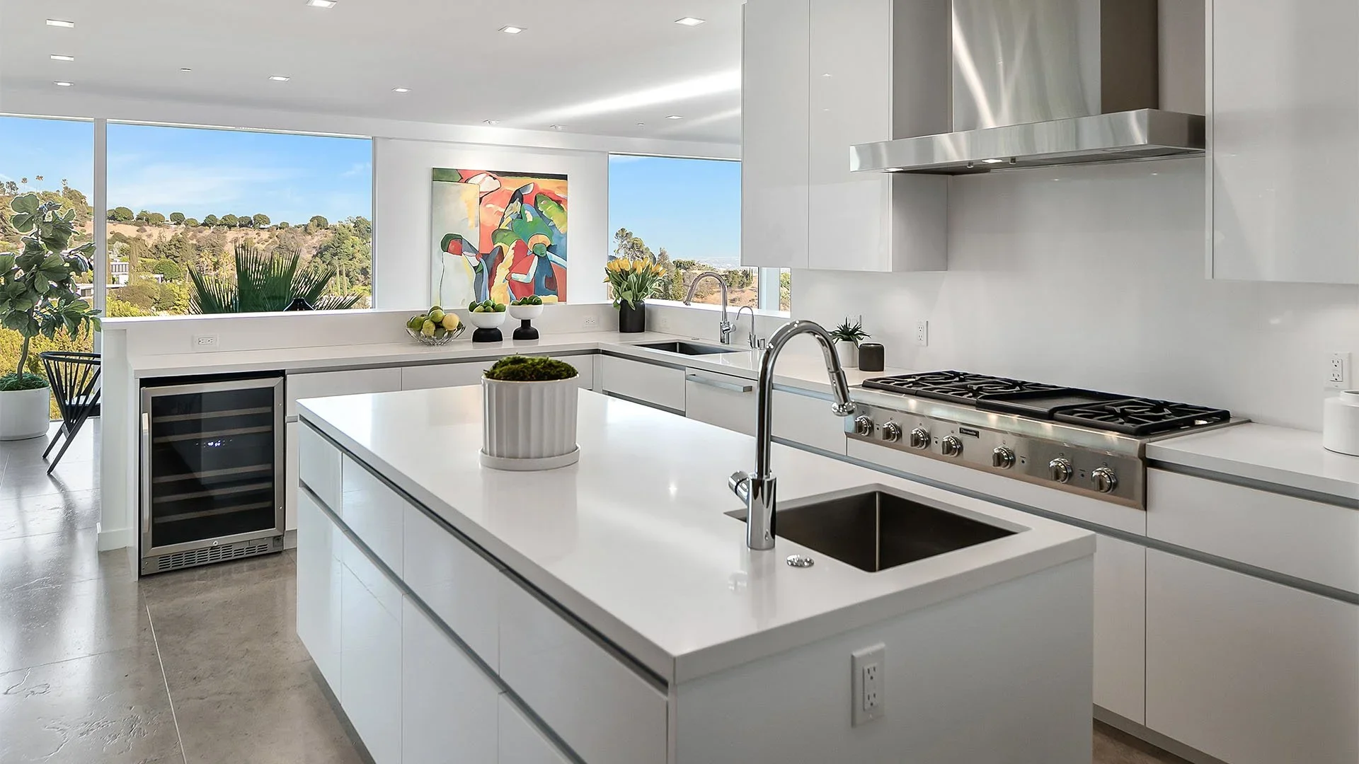 Modern kitchen with white cabinets and countertops, stainless steel stove and vent, small wine refrigerator, and large windows showing an outdoor view with green trees and hills, decorated with plants and colorful artwork.