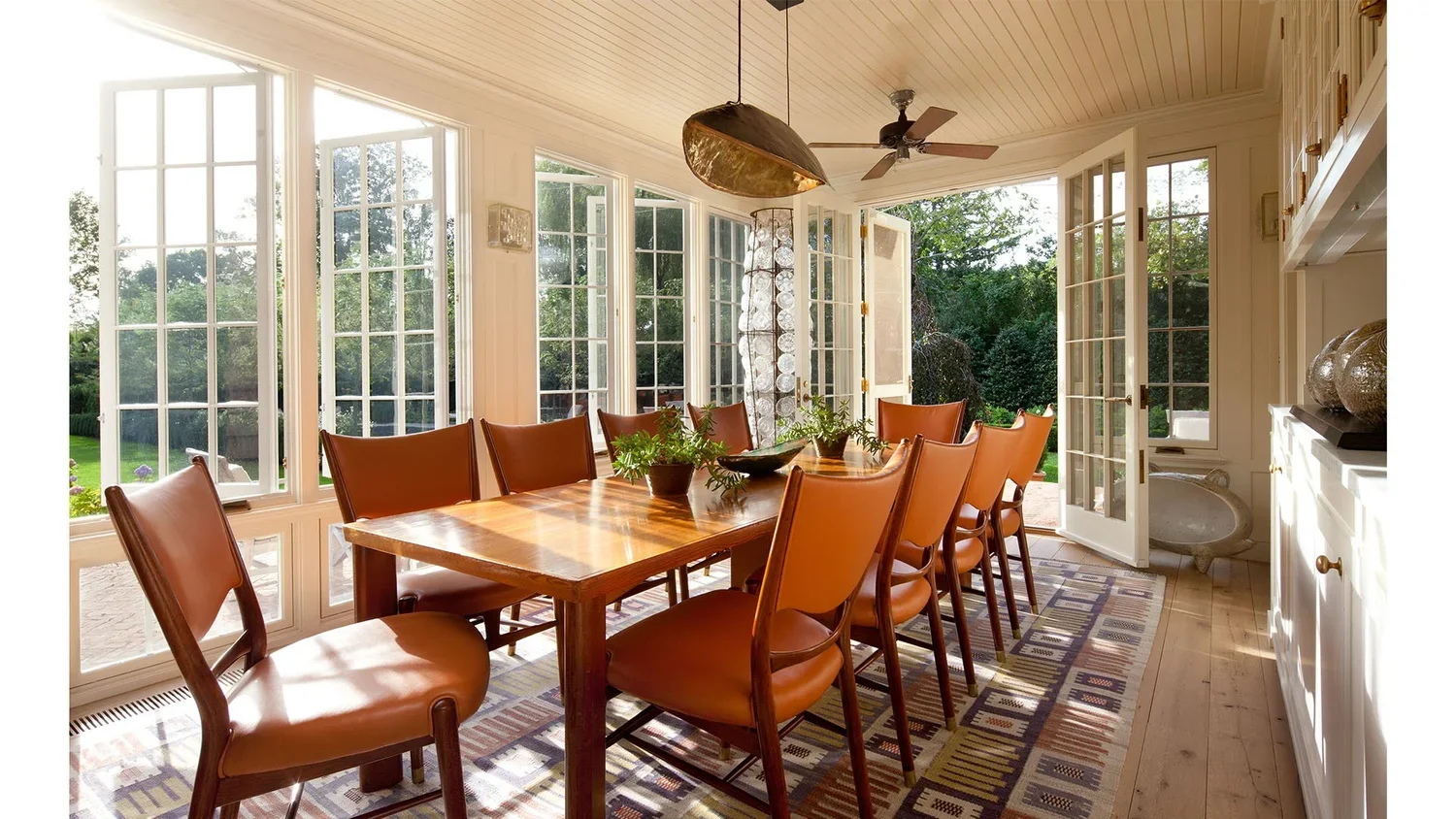 Sunlit dining room with wooden table and tan chairs, large windows and glass doors opening to a lush green outdoor space.