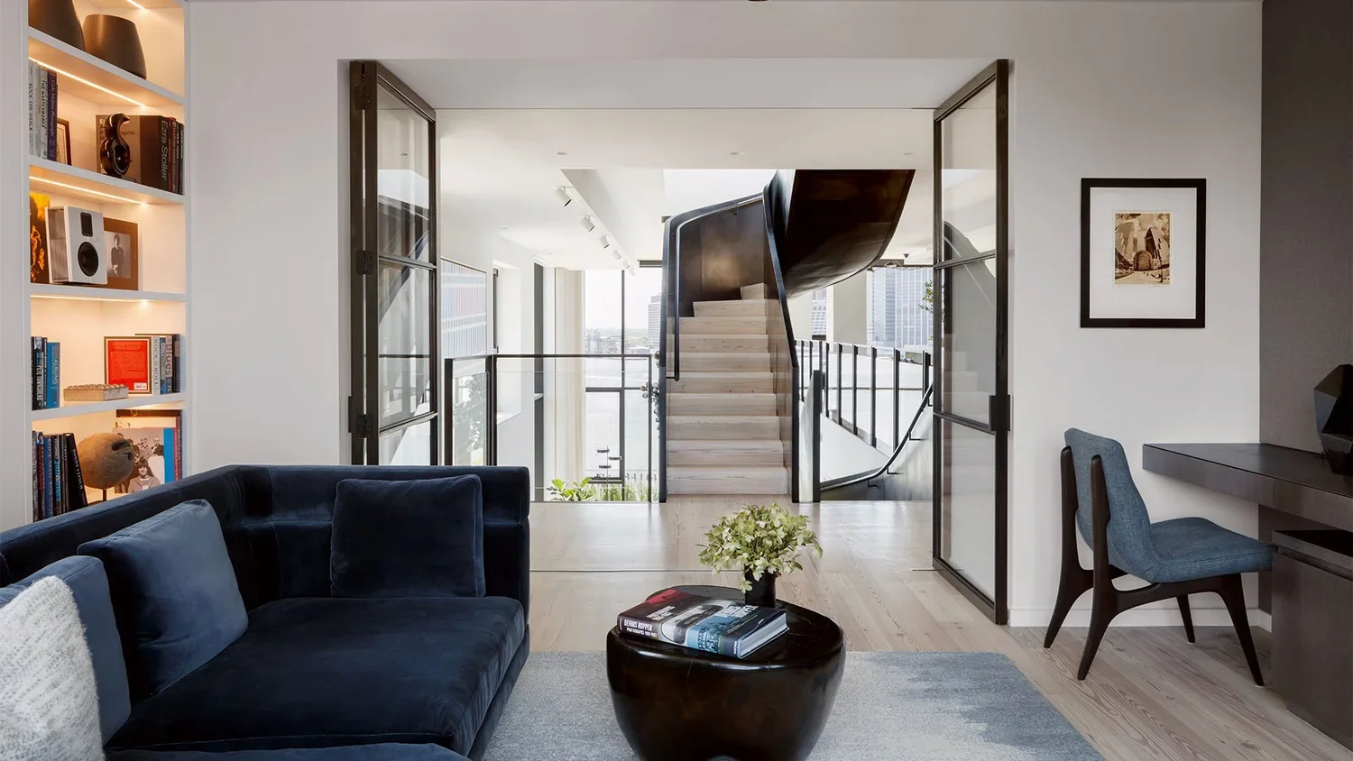 Modern living room with navy velvet sofa, coffee table with books and flowers, built-in bookshelf with books and decor, glass sliding doors, staircase with wood steps and black railing, city view in background.