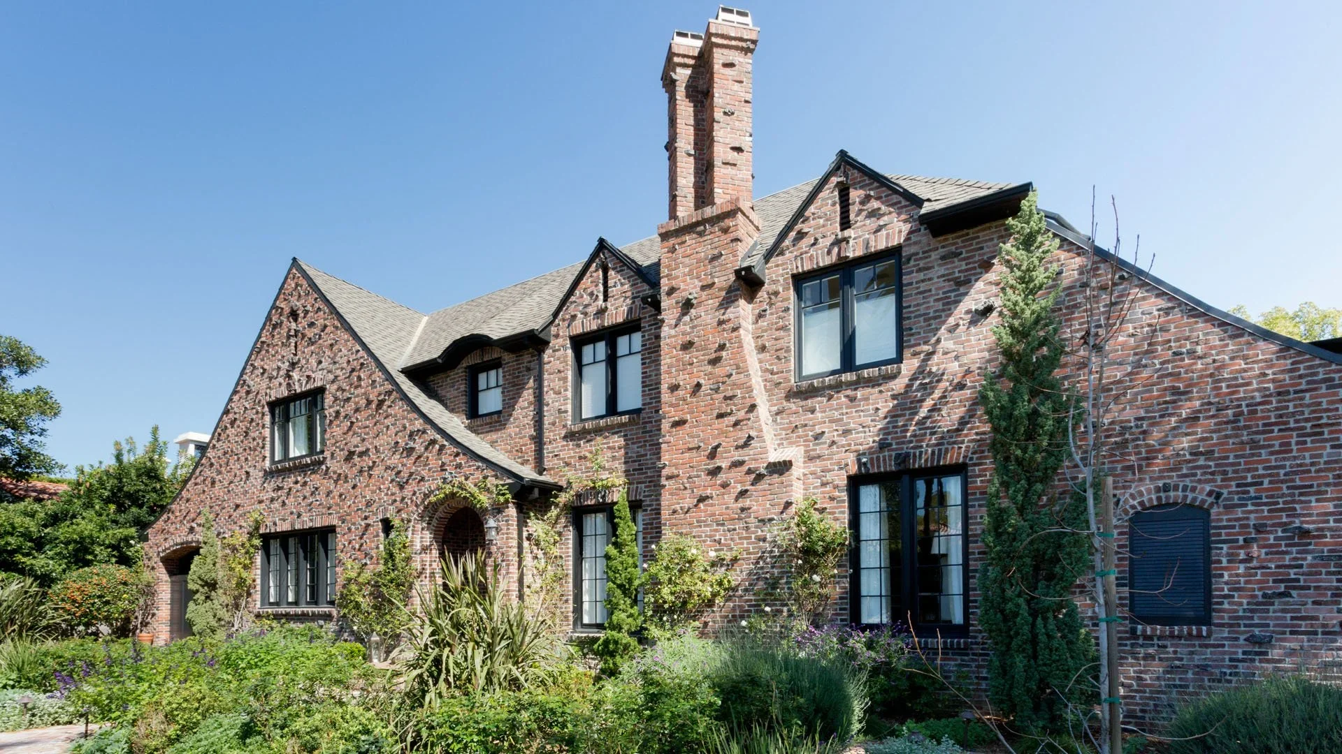Old brick house with multiple gables, black window frames, a tall brick chimney, and a landscaped garden with various plants and trees.