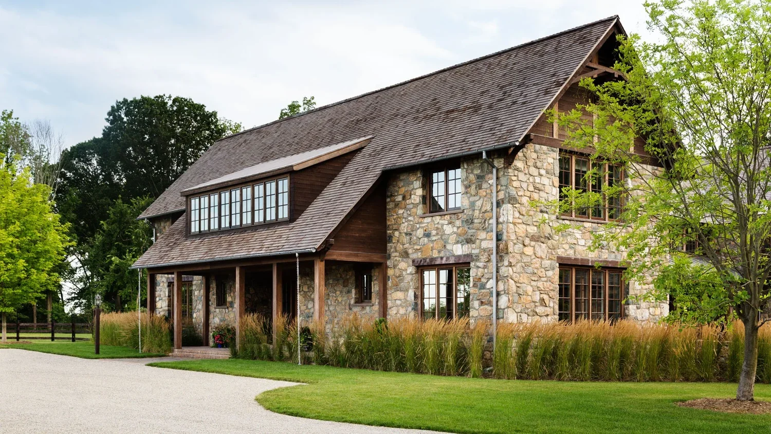 A large two-story house with a stone facade and wooden accents, surrounded by greenery and a well-kept lawn, with a curved driveway in front.