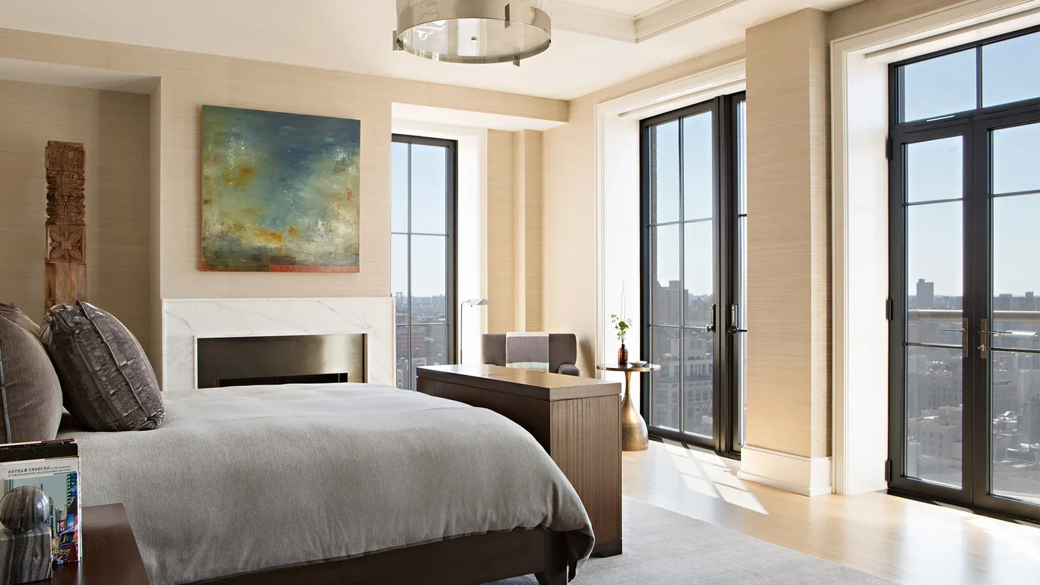 Modern bedroom with large windows, a gray bed with pillows, and artwork above the fireplace, with cityscape view outside.