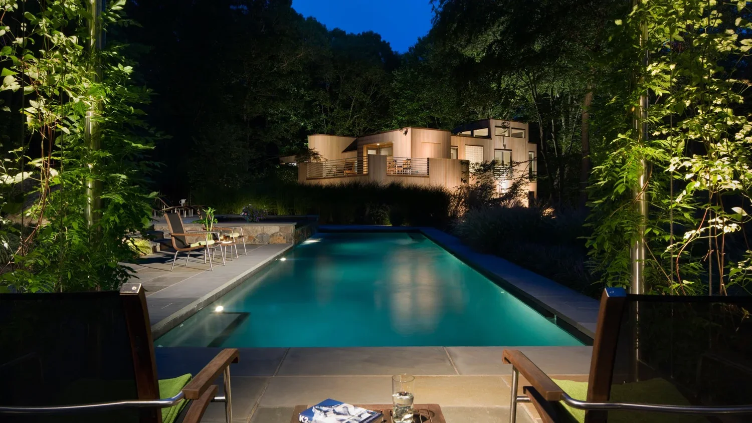Nighttime view of a modern house with illuminated pool in a lush forest setting.