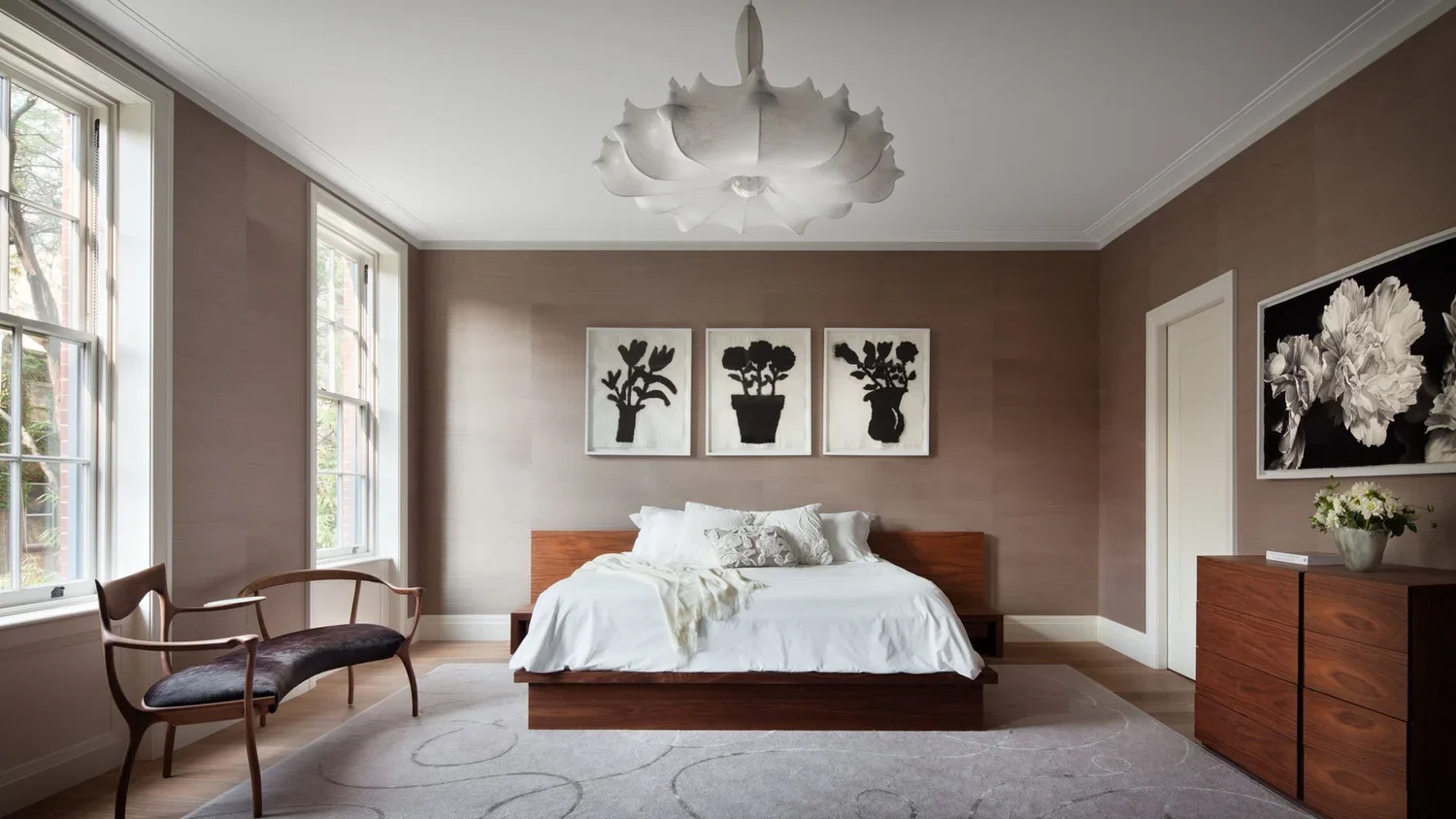 A minimalist bedroom with a wooden bed, white bedding, black and white floral artwork above the bed, a wooden dresser with a flower vase, a wooden bench with a dark cushion near the window, and large windows letting in natural light.