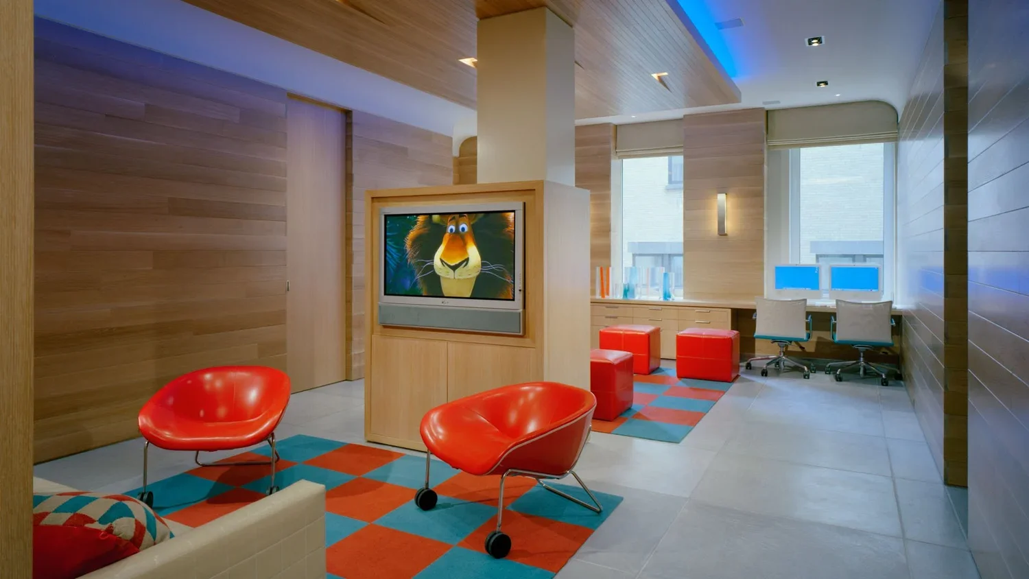 Modern hotel lobby with wooden paneling, red modern chairs, a patterned rug, a television screen showing animated lion character, and a workspace area with two computers and chairs.