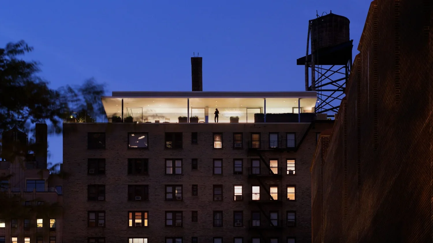 A view of a tall building at dusk with a rooftop terrace that has a person standing and taking photos, and a water tower or similar structure in the background.