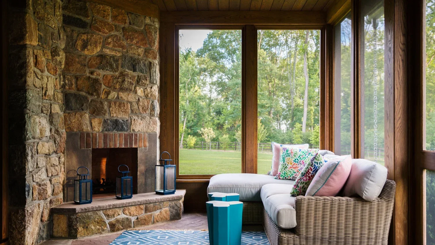Sunlit enclosed porch with large windows, wicker sectional sofa with colorful cushions, stone fireplace with lanterns on hearth, blue and white patterned rug, and scenic view of green trees and grass outside.