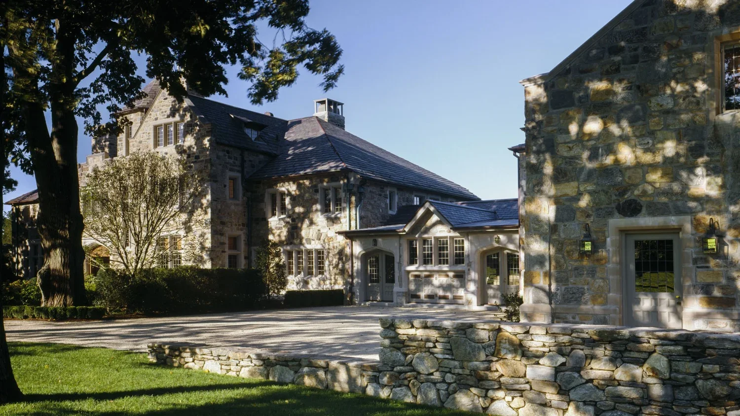 Stone mansion with multiple windows, surrounded by trees and a lawn with a stone wall in the foreground.