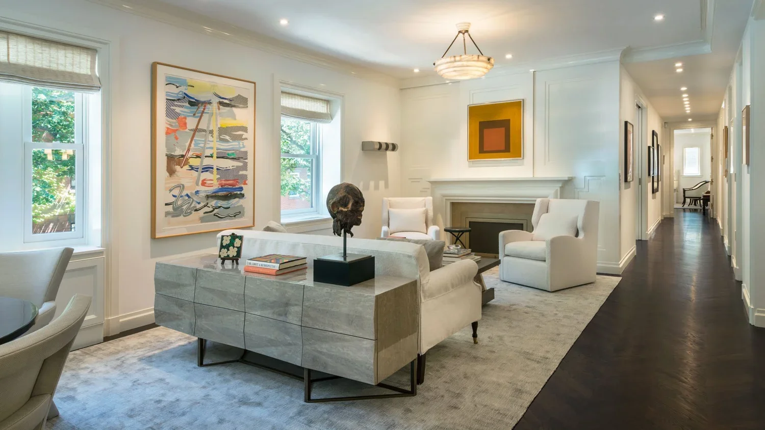 Living room with white walls, dark hardwood floors, white armchairs, a gray marble sideboard with books and a sculpture, and framed artwork on the walls.