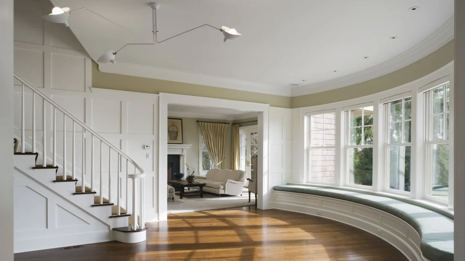 Bright living room with large bay windows, wooden floor, and built-in window seat. An adjacent room has a white sofa, fireplace, and curtains.