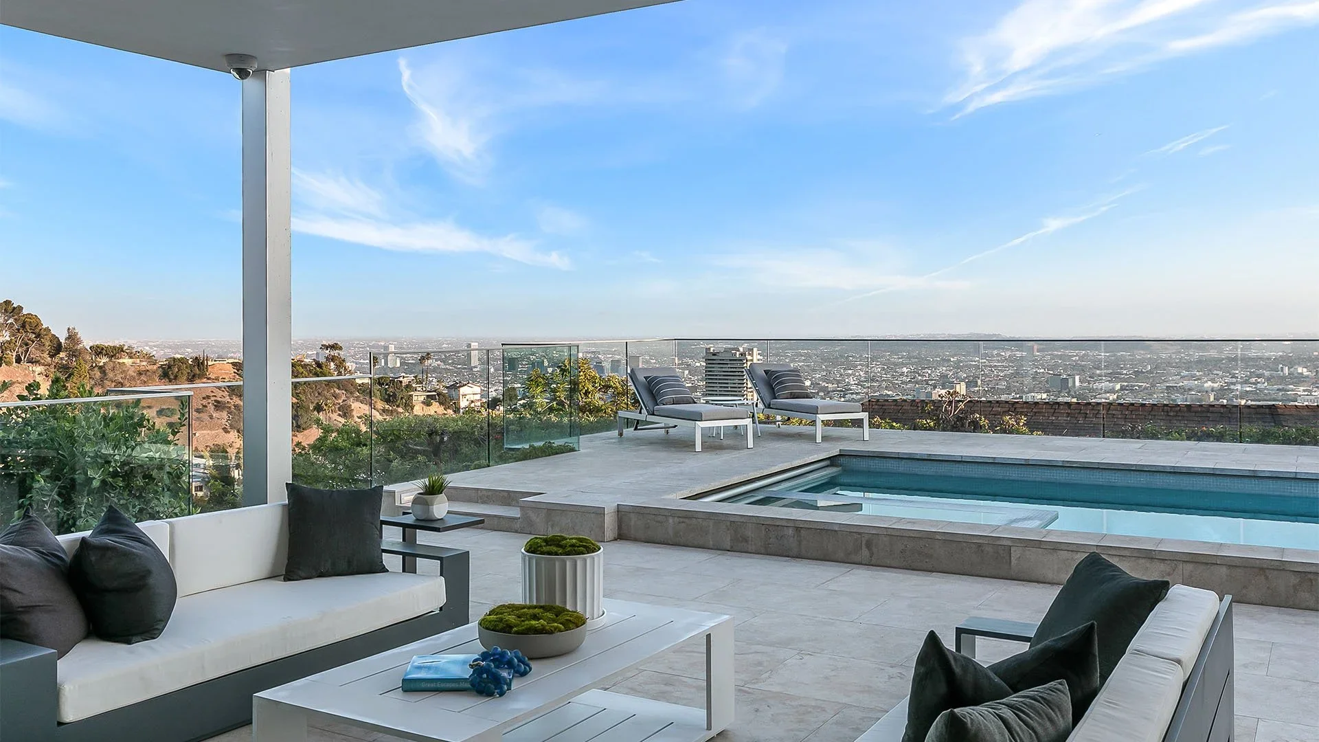 Modern outdoor terrace with a swimming pool, lounge chairs, and a city view under a blue sky with scattered clouds.