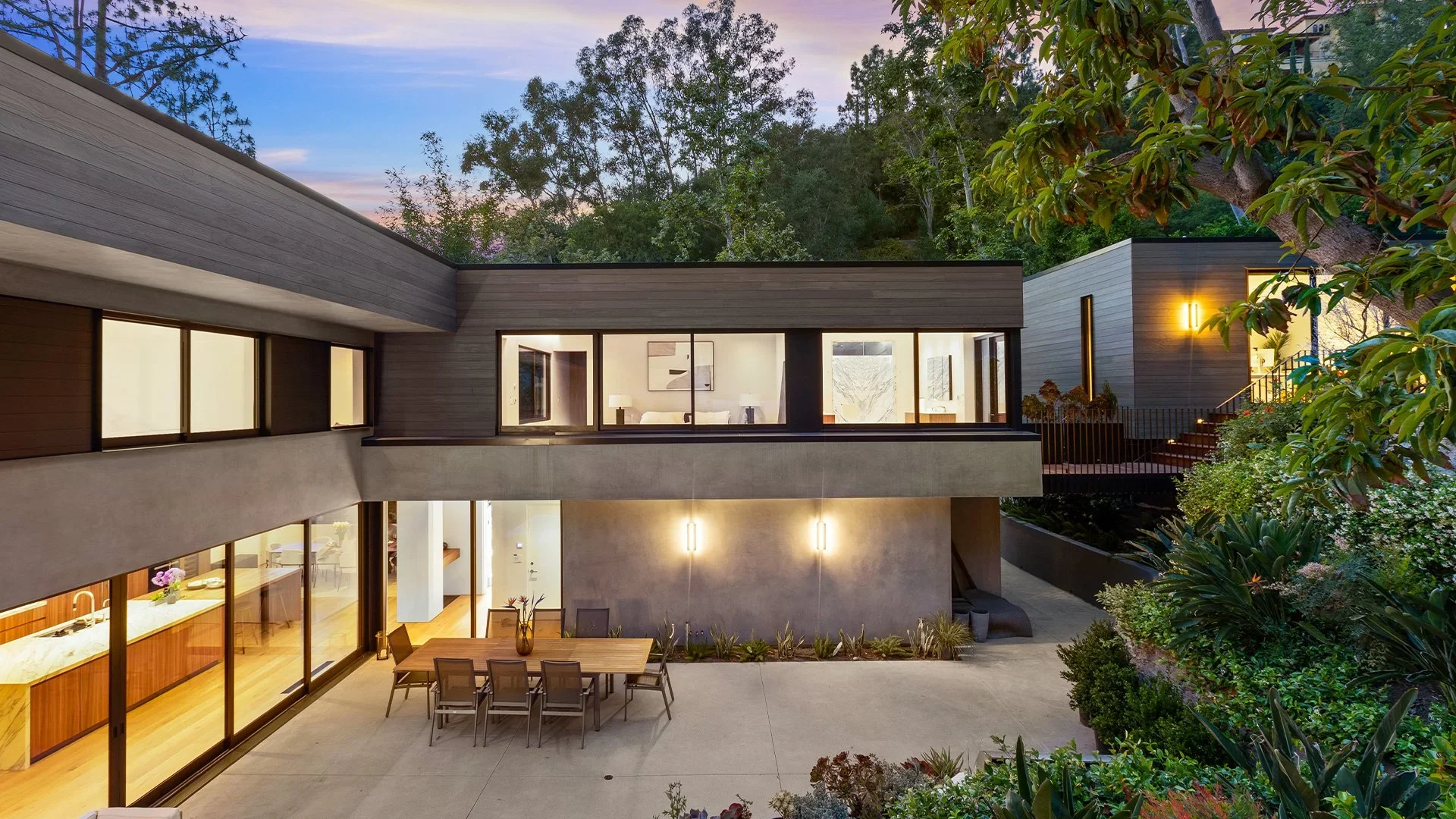 Modern multi-level house with large glass windows, outdoor patio with dining table, lush greenery, and trees in the background during evening time.