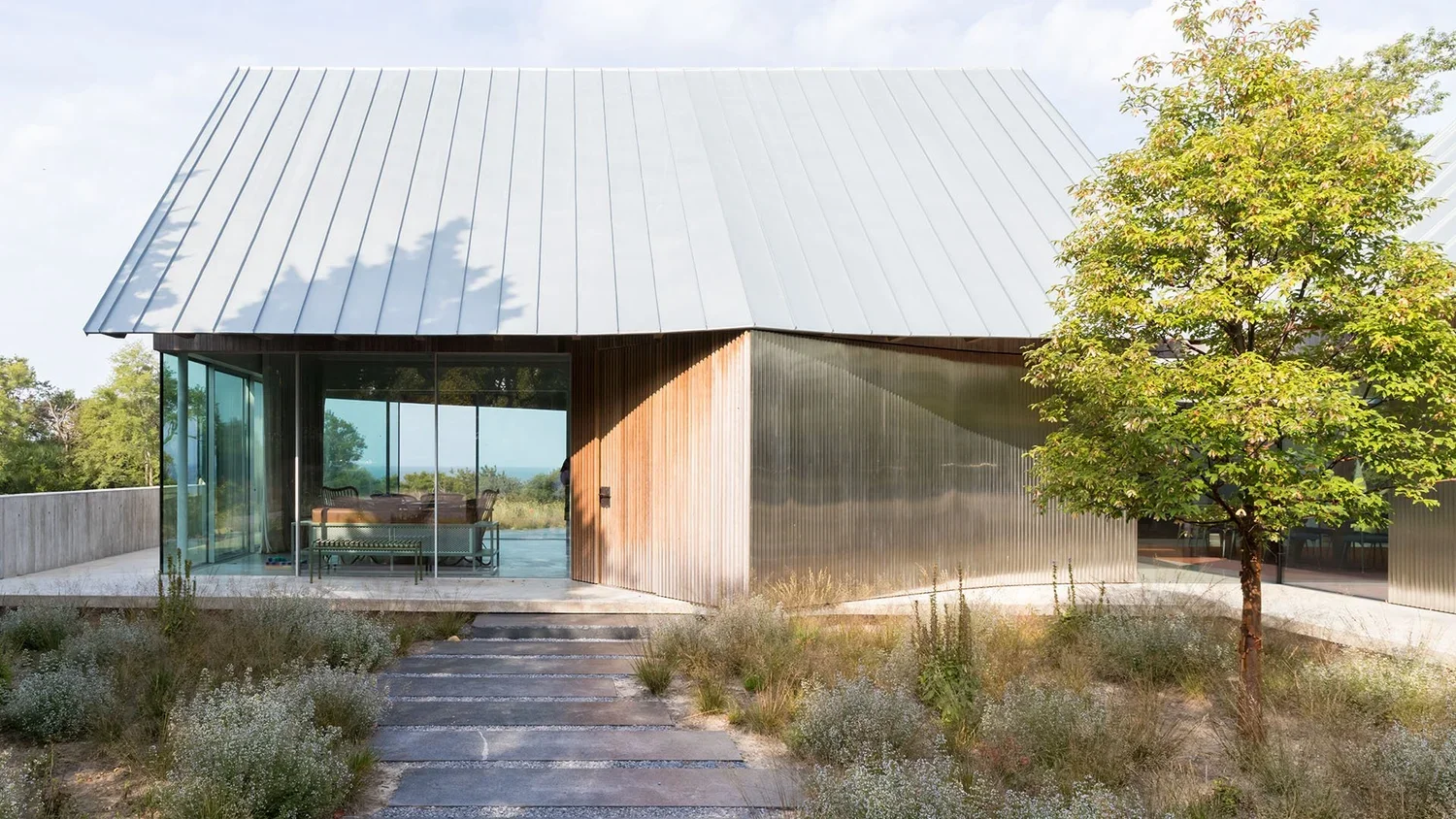 Modern house with a sloped metal roof, glass walls, and a small outdoor patio area, surrounded by landscaping and trees.