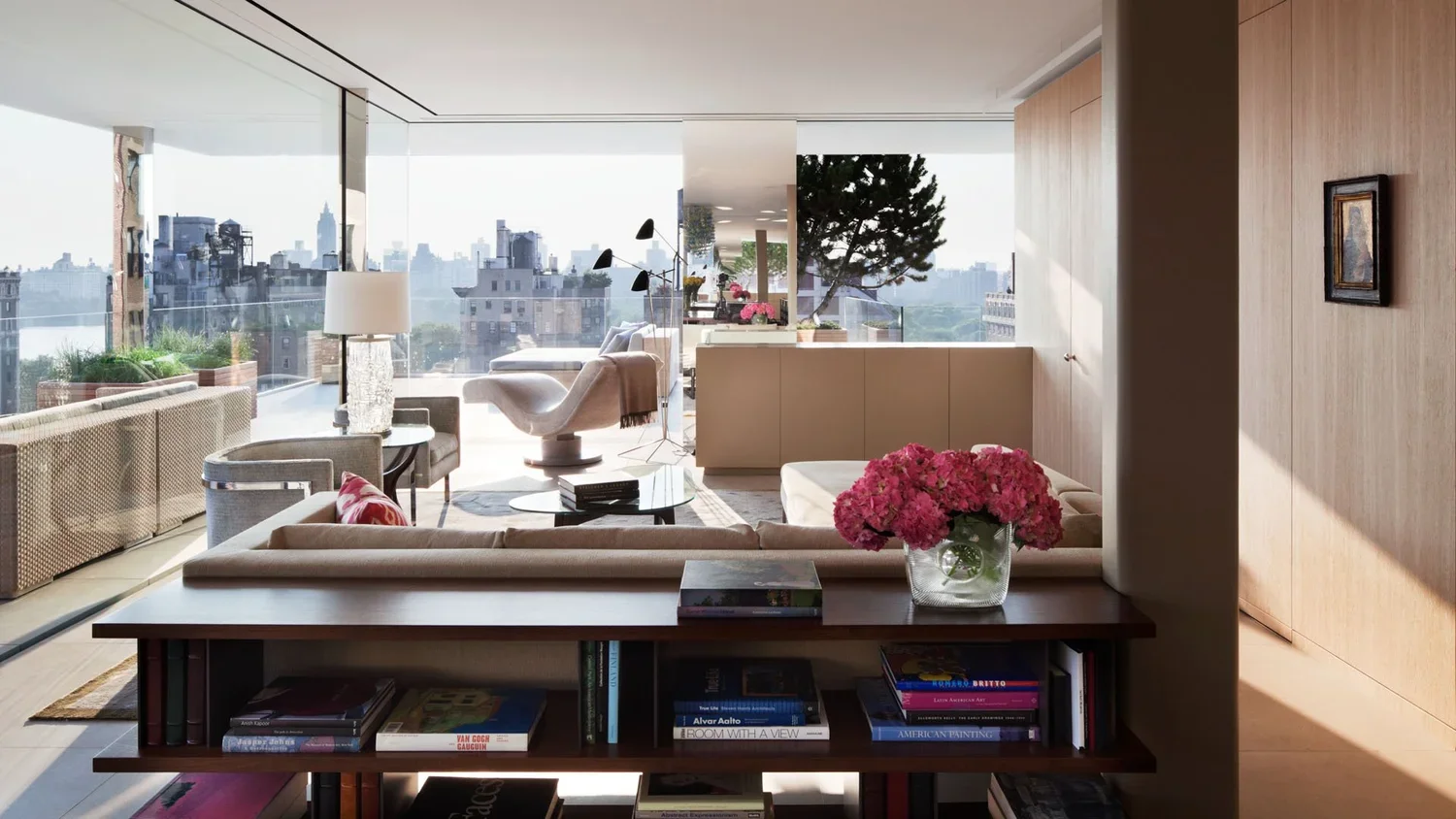 Modern living room with large windows, light-colored furniture, a coffee table, bookshelves, and pink flowers.