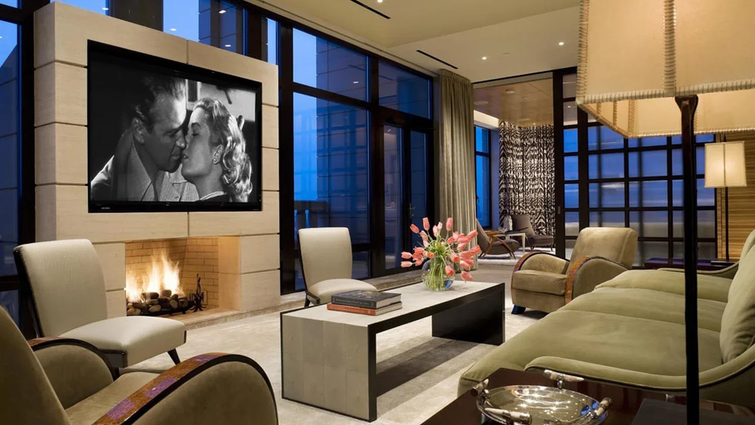 Elegant hotel lobby with large windows, modern furniture, a fireplace, and a flat-screen TV showing a black-and-white movie scene.