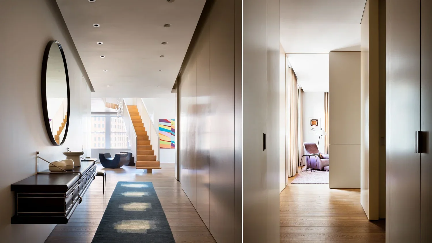 A spacious hallway with a bench and a mirror on the left, a staircase at the end, and an art piece on the wall. A doorway on the right leads to a bright living room with curtains, a chair, and artwork.