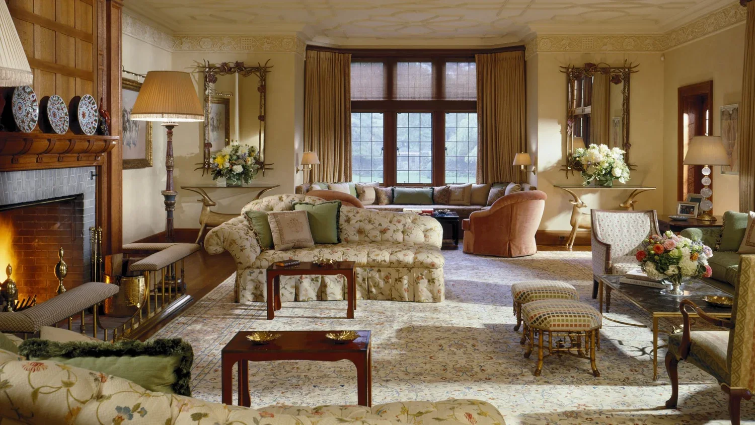 A cozy living room with floral and pastel-colored furniture, a fireplace, large windows, and various decorative items.